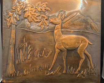 May include: A copper relief sculpture depicting a deer reaching for leaves on a tree branch. The artwork features a landscape with mountains in the background and tall grass in the foreground. The color is a warm, reddish-brown.