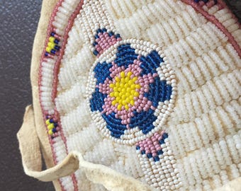 May include: A round beaded bag with a white background and a colorful floral design. The bag has a leather strap and is decorated with pink, blue, and yellow beads.