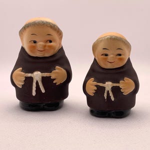 May include: Two ceramic monk figurines. The figures have smiling faces, brown robes, and white rope belts. The figurines are tan, brown, and black. The figures are standing on a white surface.