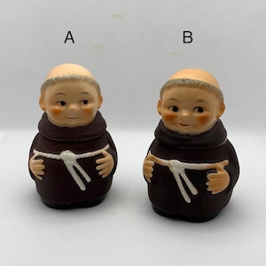 May include: Two ceramic figurines of monks, each wearing a brown robe with a white rope belt. The monks have smiling faces and are holding their hands in a prayer position. The figurines are labeled A and B.