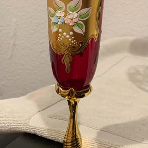 May include: A ruby red and gold-trimmed champagne flute. The glass features hand-painted floral designs in white, blue, and pink. The stem and base are gold-colored, adding to the elegant aesthetic.