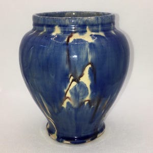 May include: A blue ceramic vase with a wide mouth and a rounded body. The vase has a glossy finish with streaks of cream and brown. The vase is a decorative item.