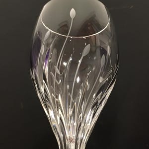 May include: A clear crystal wine glass with a long stem. The glass features etched floral designs and a rounded bowl. The etched design includes thin, curved lines and small, oval shapes. The glass is on a black surface.