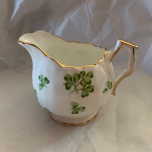 May include: A white creamer with a gold rim and green shamrock pattern. The creamer has a small handle and a rounded body.