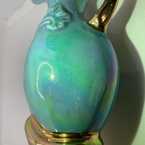 May include: A teal-colored ceramic pitcher with a gold handle and base. The pitcher has a pearlescent finish that reflects green and blue hues. A decorative leaf design is on the side near the handle.