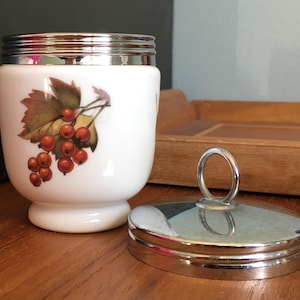 May include: White ceramic egg cup with a silver lid and a red berry design. The lid has a silver ring handle.