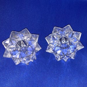 May include: Two clear, star-shaped candle holders. Each holder has a central well for a candle and is surrounded by pointed facets. The candle holders are made of a clear, faceted glass, and sit on a blue background.