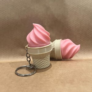 Könnte beinhalten: Zwei Eiswaffel-Schlüsselanhänger. Einer steht aufrecht, der andere liegt auf der Seite. Jeder hat eine rosa Eiscreme-Wirbel auf einem weißen Kegel mit Waffelmuster. Ein silberner Schlüsselring ist an einem befestigt.