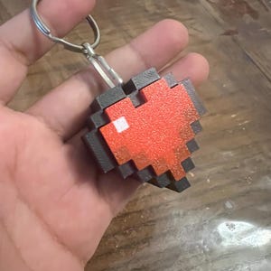 Könnte beinhalten: Pixeliger Herz-Schlüsselanhänger in Rot und Grau. Das Herzdesign hat ein weißes Quadrat in der Mitte. Der Schlüsselanhänger ist an einem silbernen Metallring befestigt.