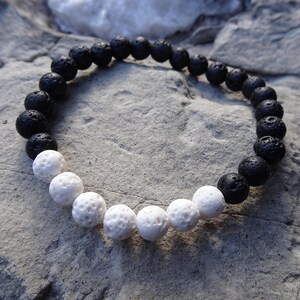 May include: A bracelet made of black and white spherical beads. The black beads have a textured, porous surface, and the white beads are smooth. The bracelet is presented on a grey, textured surface, creating a simple, natural look.