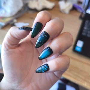 Op de afbeelding: Een hand met lange, puntige kunstnagels. De nagels zijn geverfd met een donkergroen en blauw cat-eye effect. Sommige nagels hebben een bubbelachtig ontwerp, andere een blauw-groen patroon.