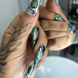 Op de afbeelding: Lange, puntige vingernagels met ingewikkelde ontwerpen. De nagels hebben een combinatie van zilver, groen en wit. Sommige nagels hebben een grote, ovale groene steen. Het ontwerp is futuristisch en artistiek.