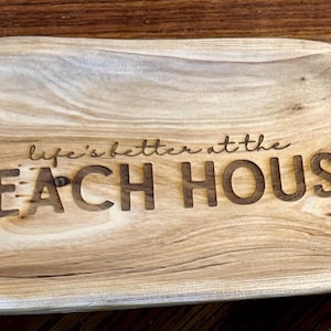 May include: A wooden serving tray with the phrase "life's better at the BEACH HOUSE" carved into the surface. The tray is oval-shaped with a light brown finish, showcasing the natural wood grain.