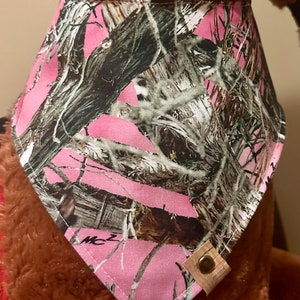 Op de afbeelding: Roze camouflage bandana met een bruine leren knoop. De bandana is gemaakt van stof en heeft een driehoekige vorm.