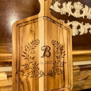 Könnte beinhalten: Holzschneidebrett mit Griff, verziert mit einem floralen Kranz und dem Buchstaben "B" in der Mitte. Der Text "Est. 2025" ist darunter eingraviert. Das Brett ist rechteckig mit einer Nut am Rand. Helle Holzfarbe.