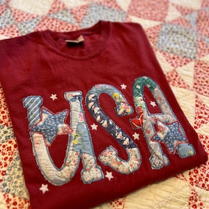 May include: A maroon crew neck t-shirt featuring the letters USA appliqued on the front. The letters are constructed from various blue, white, and red floral and patterned fabric scraps with visible stitching. Small white fabric stars are scattered around.