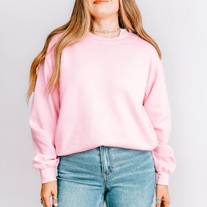 K&ouml;nnte beinhalten: Ein hellrosa Sweatshirt mit Rundhalsausschnitt. Das Sweatshirt wird mit hellblauen Jeans kombiniert. Das Model tr&auml;gt eine goldene Halskette und Ringe. Der Hintergrund ist eine schlichte, wei&szlig;e Wand.