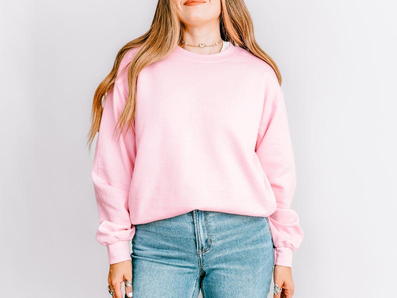K&ouml;nnte beinhalten: Ein hellrosa Sweatshirt mit Rundhalsausschnitt. Das Sweatshirt wird mit hellblauen Jeans kombiniert. Das Model tr&auml;gt eine goldene Halskette und Ringe. Der Hintergrund ist eine schlichte, wei&szlig;e Wand.