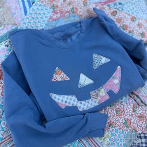 May include: A blue crewneck sweatshirt featuring a jack-o'-lantern face applique on the front. The eyes, nose, and jagged mouth are crafted from various colorful floral and checkered fabric patches, each secured with visible white stitching.