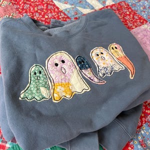 May include: A slate blue crewneck sweatshirt featuring five ghost-shaped fabric appliqués across the chest. Each ghost is crafted from different floral and patterned cotton fabrics in shades of green, pink, yellow, white, blue, and orange.