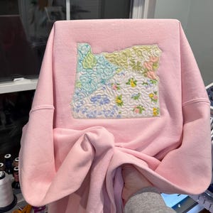 K&ouml;nnte beinhalten: Hellrosa Sweatshirt mit einem Patch des Bundesstaates Oregon. Der Patch besteht aus verschiedenen Stoffmustern in Blau-, Gr&uuml;n-, Gelb- und Rosat&ouml;nen. Das Sweatshirt hat lange &Auml;rmel und einen Rundhalsausschnitt.