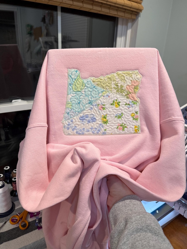 K&ouml;nnte beinhalten: Hellrosa Sweatshirt mit einem Patch des Bundesstaates Oregon. Der Patch besteht aus verschiedenen Stoffmustern in Blau-, Gr&uuml;n-, Gelb- und Rosat&ouml;nen. Das Sweatshirt hat lange &Auml;rmel und einen Rundhalsausschnitt.
