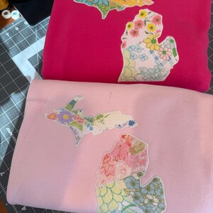 K&ouml;nnte beinhalten: Zwei gefaltete Sweatshirts, eines in Pink und eines in Hellrosa. Jedes hat ein florales Patchwork-Design in Form des Bundesstaates Michigan. Das Patchwork umfasst verschiedene Blumenmuster in Rosa-, Gelb-, Blau- und Gr&uuml;nt&ouml;nen.