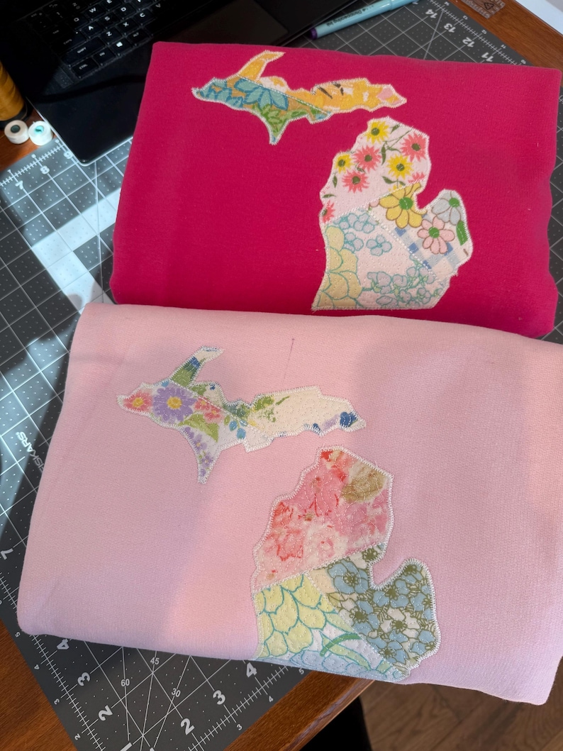 K&ouml;nnte beinhalten: Zwei gefaltete Sweatshirts, eines in Pink und eines in Hellrosa. Jedes hat ein florales Patchwork-Design in Form des Bundesstaates Michigan. Das Patchwork umfasst verschiedene Blumenmuster in Rosa-, Gelb-, Blau- und Gr&uuml;nt&ouml;nen.