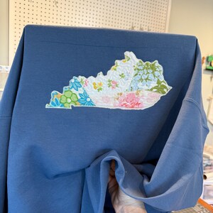 K&ouml;nnte beinhalten: Blaues Sweatshirt mit einem gestickten Kentucky-Aufn&auml;her. Der Aufn&auml;her besteht aus floralen Stoffen in Rosa, Blau, Gr&uuml;n und Wei&szlig;. Das Sweatshirt hat eine einfarbige blaue Farbe. Der Aufn&auml;her hat die Form des Bundesstaates Kentucky.