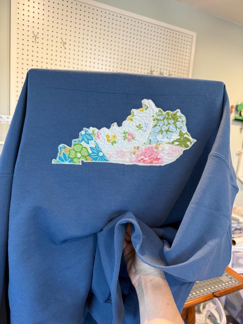 K&ouml;nnte beinhalten: Blaues Sweatshirt mit einem gestickten Kentucky-Aufn&auml;her. Der Aufn&auml;her besteht aus floralen Stoffen in Rosa, Blau, Gr&uuml;n und Wei&szlig;. Das Sweatshirt hat eine einfarbige blaue Farbe. Der Aufn&auml;her hat die Form des Bundesstaates Kentucky.