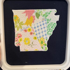 K&ouml;nnte beinhalten: Ein Patchwork-Flicken in Form des US-Bundesstaates Arkansas, mit einem Patchwork-Design mit floralen und geometrischen Mustern in verschiedenen Farben. Der Flicken befindet sich auf einem dunkelblauen Hintergrund und wird in einem Stickrahmen gehalten. Der Rahmen misst 31.1 cm x 28.8 cm.