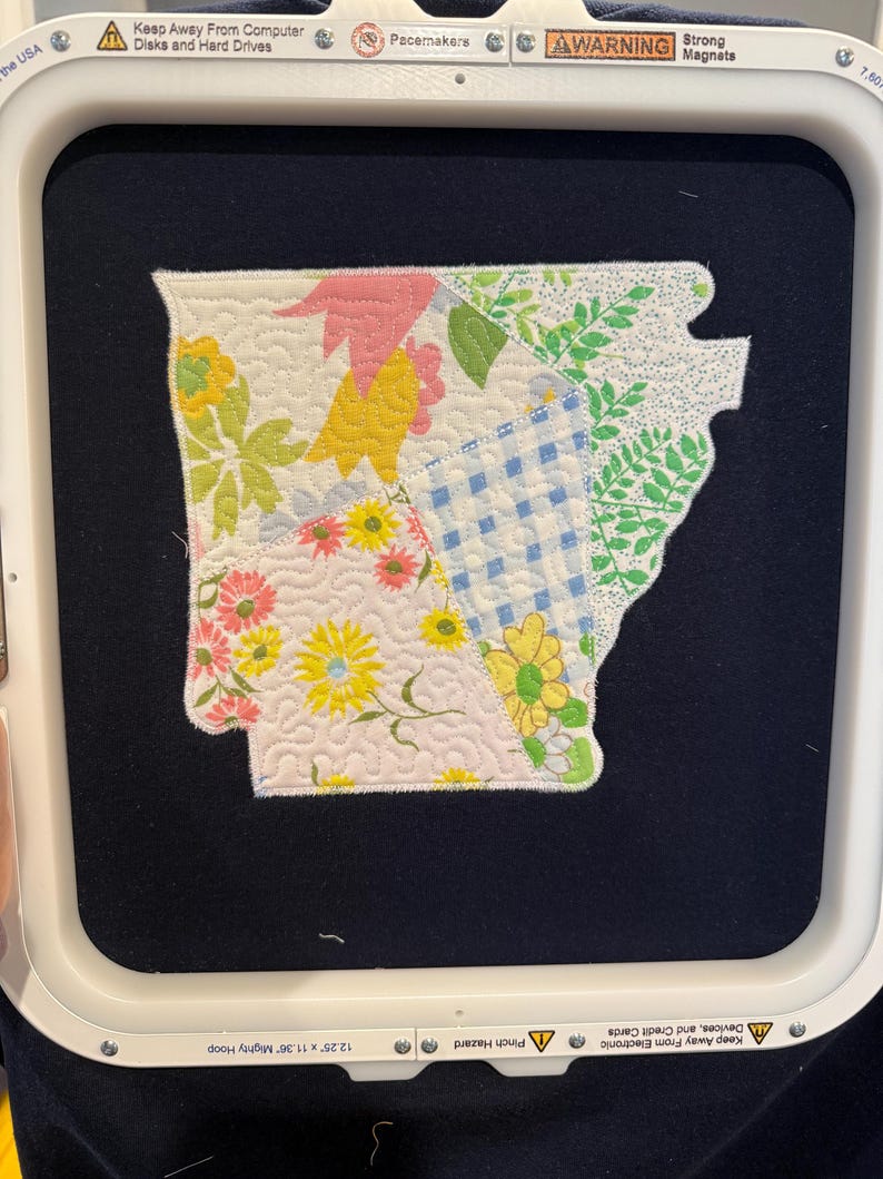 K&ouml;nnte beinhalten: Ein Patchwork-Flicken in Form des US-Bundesstaates Arkansas, mit einem Patchwork-Design mit floralen und geometrischen Mustern in verschiedenen Farben. Der Flicken befindet sich auf einem dunkelblauen Hintergrund und wird in einem Stickrahmen gehalten. Der Rahmen misst 31.1 cm x 28.8 cm.