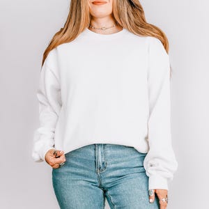 K&ouml;nnte beinhalten: Ein wei&szlig;es Sweatshirt mit Rundhalsausschnitt und langen &Auml;rmeln. Das Sweatshirt wird mit hellblauen Jeans kombiniert. Das Model tr&auml;gt eine Halskette und Ringe. Der Hintergrund ist eine schlichte wei&szlig;e Wand.