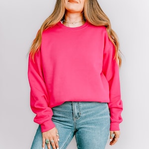 K&ouml;nnte beinhalten: Ein leuchtend pinkfarbener Pullover mit langen &Auml;rmeln. Der Pullover wird mit hellblauen Jeans kombiniert. Das Model tr&auml;gt eine Halskette und Ringe. Der Hintergrund ist eine schlichte wei&szlig;e Wand.