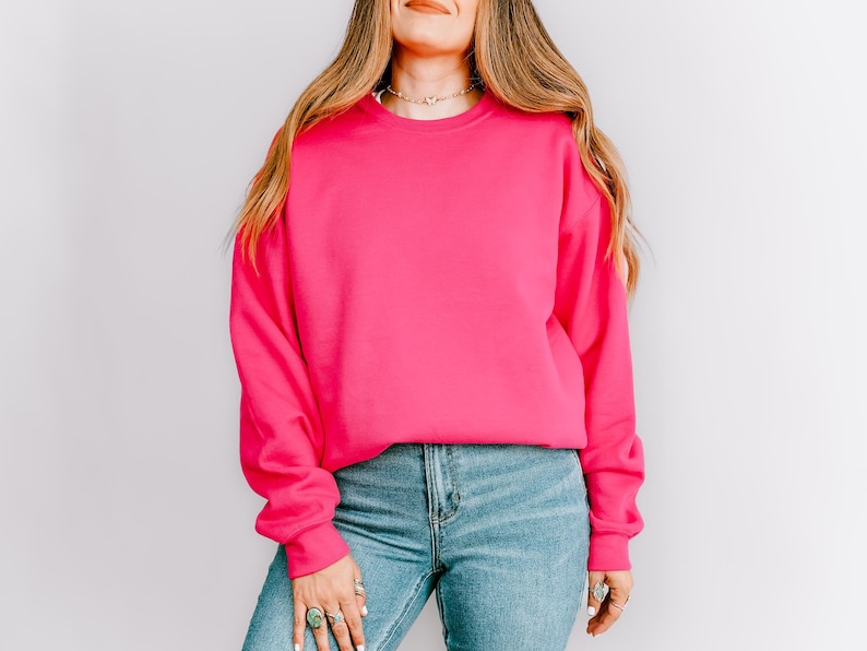 K&ouml;nnte beinhalten: Ein leuchtend pinkfarbener Pullover mit langen &Auml;rmeln. Der Pullover wird mit hellblauen Jeans kombiniert. Das Model tr&auml;gt eine Halskette und Ringe. Der Hintergrund ist eine schlichte wei&szlig;e Wand.