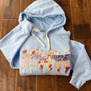 May include: A light blue hooded sweatshirt with a design of five colorful patchwork bows on the front.