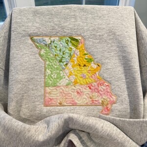 K&ouml;nnte beinhalten: Ein graues Sweatshirt mit einem farbenfrohen, gesteppten Design des Bundesstaates Missouri. Der Umriss von Missouri ist mit floralen Mustern in Gr&uuml;n-, Gelb- und Rosat&ouml;nen gef&uuml;llt, die mit goldenen N&auml;hten umrandet sind.