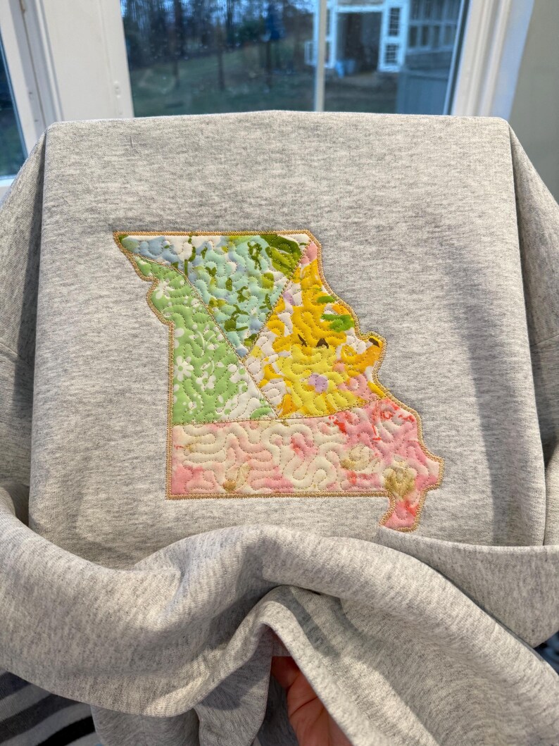 K&ouml;nnte beinhalten: Ein graues Sweatshirt mit einem farbenfrohen, gesteppten Design des Bundesstaates Missouri. Der Umriss von Missouri ist mit floralen Mustern in Gr&uuml;n-, Gelb- und Rosat&ouml;nen gef&uuml;llt, die mit goldenen N&auml;hten umrandet sind.