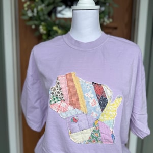 May include: A light purple t-shirt featuring a patchwork design in the shape of the state of Wisconsin. The patchwork includes various fabric patterns and colors, including red, blue, and yellow. The shirt is displayed on a mannequin.