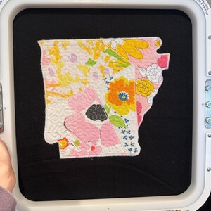 K&ouml;nnte beinhalten: Ein farbenfroher, gesteppter Aufn&auml;her in Form des Bundesstaates Arkansas mit floralen und geometrischen Mustern in Gelb, Rosa, Orange und Wei&szlig;. Der Aufn&auml;her wird in einem 31.1 cm x 28.8 cm gro&szlig;en Stickrahmen gehalten. Der Rahmen hat Sicherheitshinweise und Text.