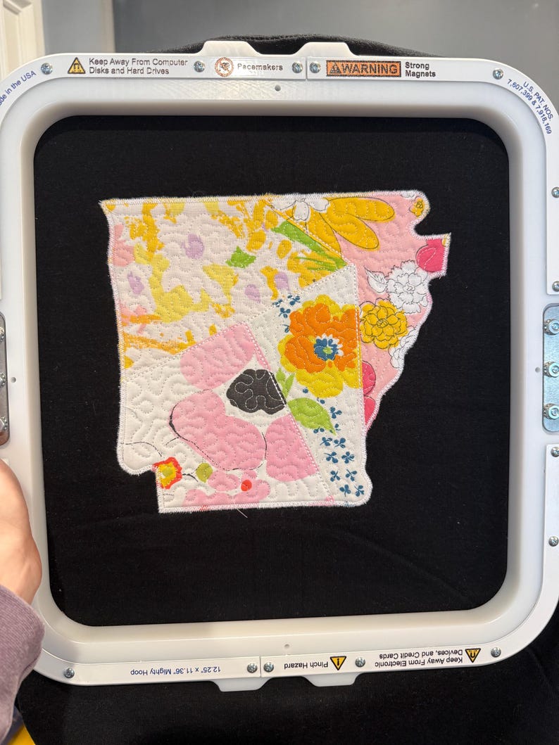 K&ouml;nnte beinhalten: Ein farbenfroher, gesteppter Aufn&auml;her in Form des Bundesstaates Arkansas mit floralen und geometrischen Mustern in Gelb, Rosa, Orange und Wei&szlig;. Der Aufn&auml;her wird in einem 31.1 cm x 28.8 cm gro&szlig;en Stickrahmen gehalten. Der Rahmen hat Sicherheitshinweise und Text.