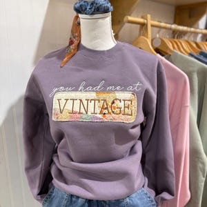 May include: A mauve long-sleeve sweatshirt featuring the embroidered white text you had me at and a rectangular patch with a multicolored floral quilt pattern containing the brown embroidered word VINTAGE on the front center.