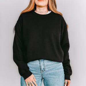 K&ouml;nnte beinhalten: Ein schwarzes Sweatshirt mit Rundhalsausschnitt und langen &Auml;rmeln. Das Sweatshirt wird mit hellblauen Jeans kombiniert. Das Model tr&auml;gt ein wei&szlig;es Unterhemd und eine goldene Halskette. Der Hintergrund ist eine schlichte wei&szlig;e Wand.