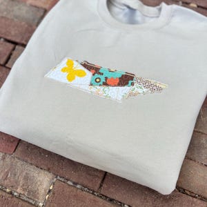 K&ouml;nnte beinhalten: Beiges Sweatshirt mit einem aufgestickten Aufn&auml;her in Form eines Staates. Der Aufn&auml;her enth&auml;lt einen gelben Schmetterling, florale Muster und verschiedene Farben. Das Sweatshirt liegt gefaltet auf einer Ziegeloberfl&auml;che.