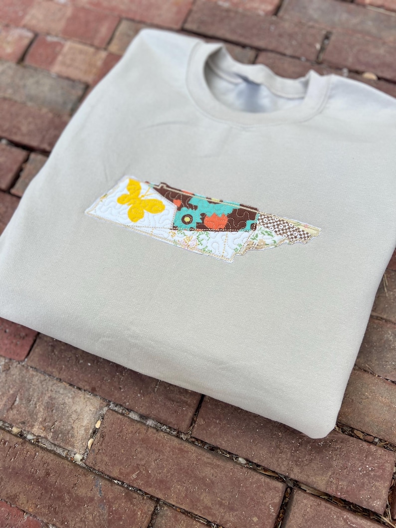 K&ouml;nnte beinhalten: Beiges Sweatshirt mit einem aufgestickten Aufn&auml;her in Form eines Staates. Der Aufn&auml;her enth&auml;lt einen gelben Schmetterling, florale Muster und verschiedene Farben. Das Sweatshirt liegt gefaltet auf einer Ziegeloberfl&auml;che.