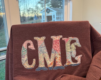 Könnte beinhalten: Ein kastanienbraunes Sweatshirt mit den aufgenähten Buchstaben CME auf der Vorderseite. Die Buchstaben sind mit einem Patchwork aus verschiedenen Blumen- und Vichy-Stoffmustern in Rosa, Blau, Gelb und Grün gefüllt und mit sichtbaren Nähten umrandet.