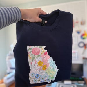 K&ouml;nnte beinhalten: Marineblaues Sweatshirt mit einem floralen Patchwork-Design in Form des Bundesstaates Georgia. Das Patchwork umfasst rosa Rosen, gelbe Blumen und blaue G&auml;nsebl&uuml;mchen. Das Sweatshirt wird von einer Person gehalten.
