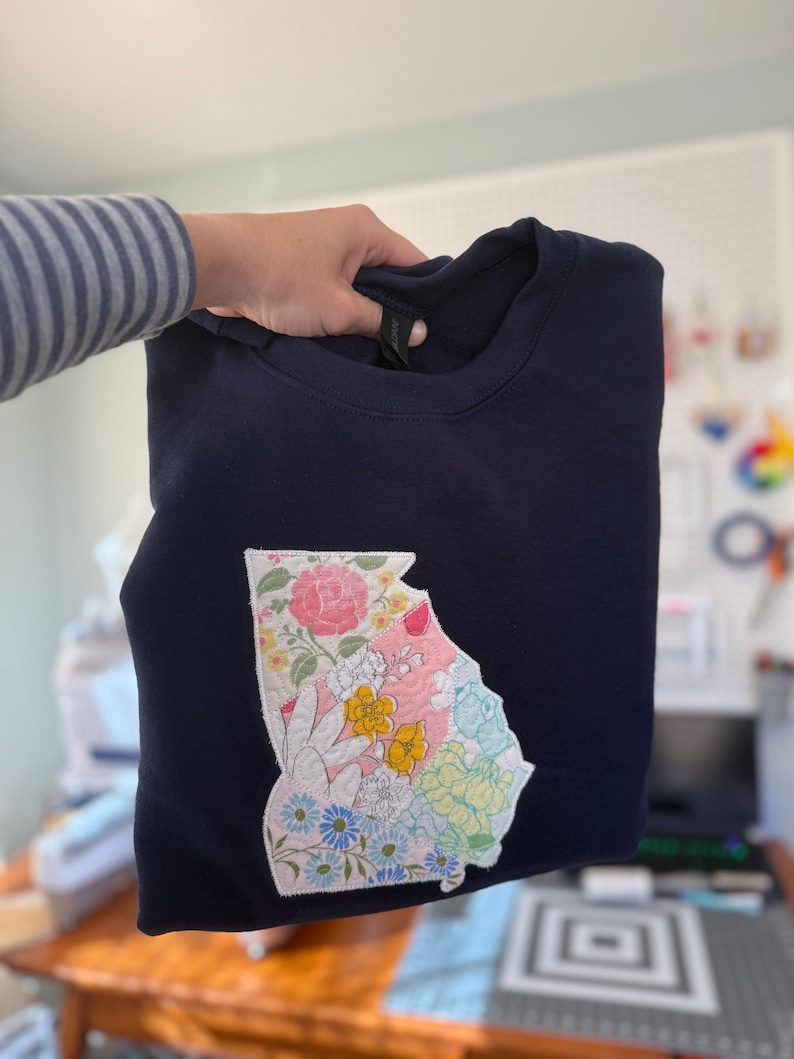 K&ouml;nnte beinhalten: Marineblaues Sweatshirt mit einem floralen Patchwork-Design in Form des Bundesstaates Georgia. Das Patchwork umfasst rosa Rosen, gelbe Blumen und blaue G&auml;nsebl&uuml;mchen. Das Sweatshirt wird von einer Person gehalten.