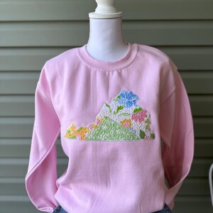 K&ouml;nnte beinhalten: Ein hellrosa Sweatshirt mit einem gestickten Design des Bundesstaates Virginia. Das Design umfasst bunte Blumen und eine gr&uuml;ne Basis, mit einer blauen Blume oben. Das Sweatshirt wird auf einer Schaufensterpuppe pr&auml;sentiert.