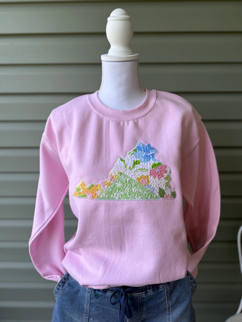 K&ouml;nnte beinhalten: Ein hellrosa Sweatshirt mit einem gestickten Design des Bundesstaates Virginia. Das Design umfasst bunte Blumen und eine gr&uuml;ne Basis, mit einer blauen Blume oben. Das Sweatshirt wird auf einer Schaufensterpuppe pr&auml;sentiert.